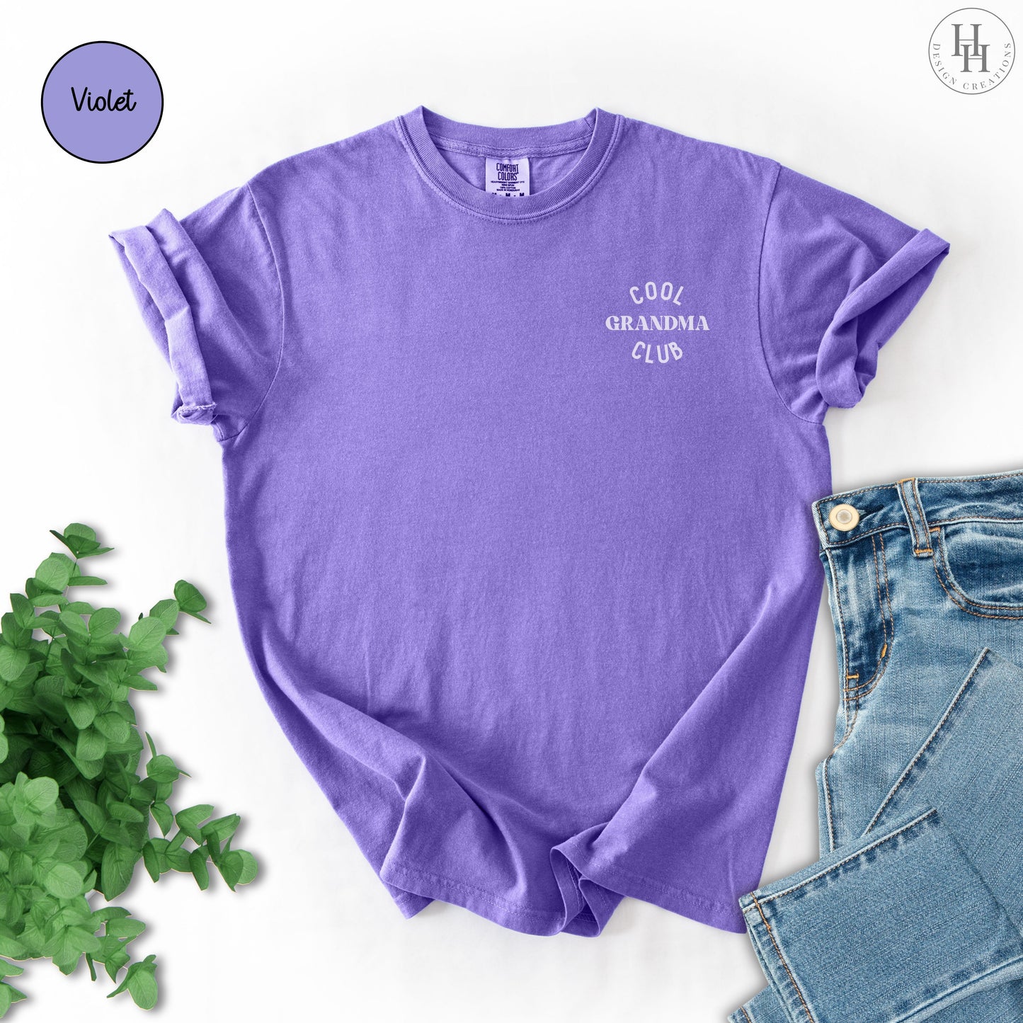 Purple t-shirt with 'Cool Grandma Club' text on a white background with jeans and greenery.
